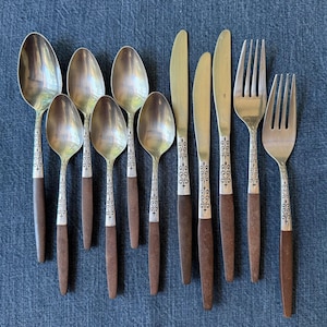 May include: A set of stainless steel flatware consisting of five spoons, two knives, and two forks. Each piece features a silver-toned metal bowl or head with an ornate scrollwork pattern on the neck and a solid brown handle at the base.
