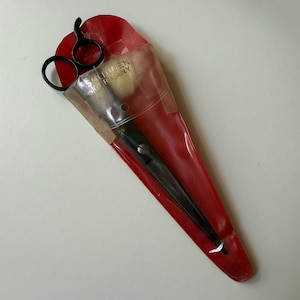 May include: Black scissors in a red plastic sheath. The scissors are partially visible, with the handles and blades extending from the sheath. The sheath has the text "SOLINGEN GERMANY" printed on it. The scissors appear to be for crafting or sewing.