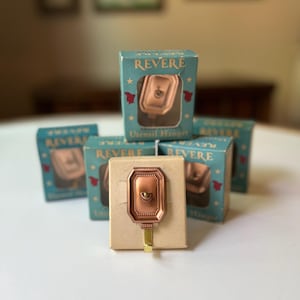 May include: A collection of vintage "Revere" utensil hangers in their original packaging. The copper-toned hangers are displayed in small, teal boxes with the product name and a graphic of the hanger. The boxes are arranged on a white surface.