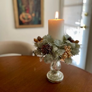 May include: A white pillar candle with a flame sits on a silver candlestick. The candlestick is decorated with a wreath of faux greenery, pine cones, and berries.