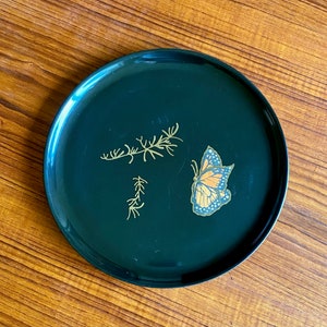 May include: A black circular tray with a gold butterfly and floral design. The butterfly has orange and black wings.