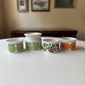 May include: Four ceramic containers with various fruit designs. Two small containers feature green apples, one has red berries and green leaves, and the last one has orange slices. The containers are white with a simple, rounded shape.