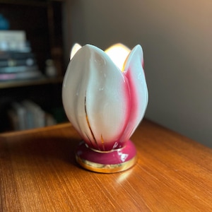 May include: A white and pink ceramic flower-shaped vase with gold accents. The vase has a wide, open top and a narrow base.