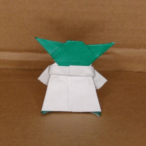 May include: A green origami Yoda figure with a white robe. The figure is standing on a brown surface.