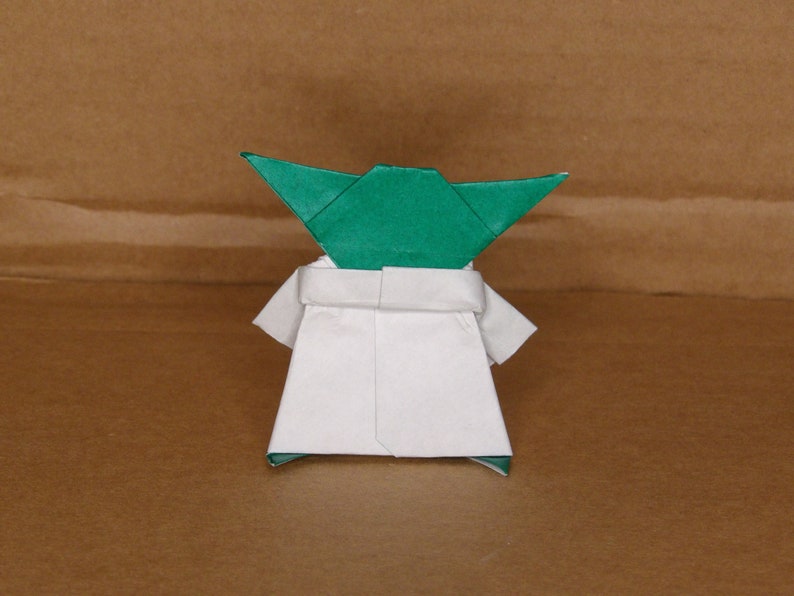 May include: A green origami Yoda figure with a white robe. The figure is standing on a brown surface.