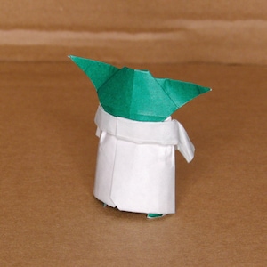 May include: A green and white origami figure of Yoda from Star Wars. The figure is folded in a traditional origami style, with a green head and a white robe.