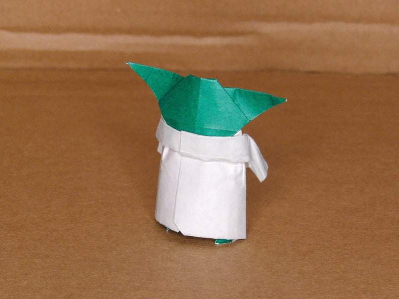 May include: A green and white origami figure of Yoda from Star Wars. The figure is folded in a traditional origami style, with a green head and a white robe.