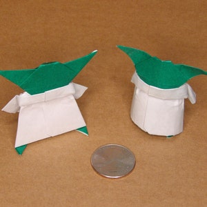 May include: Two origami Yoda figures made of green and white paper. The figures are standing on a brown surface.