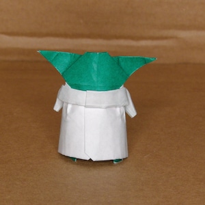 May include: A green and white origami figure of a character from the Star Wars franchise. The figure is standing on a brown surface.