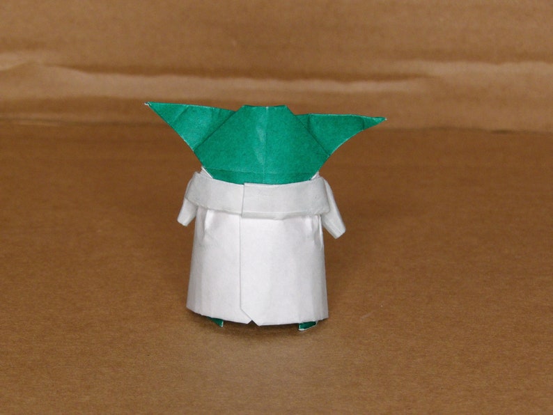 May include: A green and white origami figure of a character from the Star Wars franchise. The figure is standing on a brown surface.