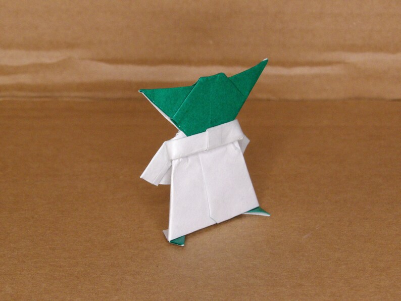 May include: Green and white origami figure of Yoda from Star Wars. The figure is standing with its arms outstretched.