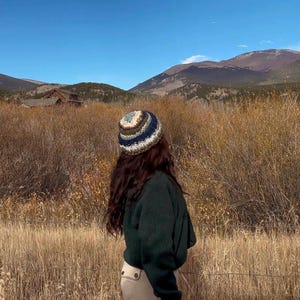 May include: A person wearing a green sweater and a colorful crocheted beanie hat stands in a field of tall grass, looking out at a mountain range in the distance.