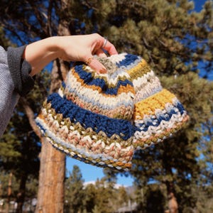 May include: Two knitted hats in a colorful striped pattern with shades of blue, yellow, white, and brown. The hats are held in a person's hand.