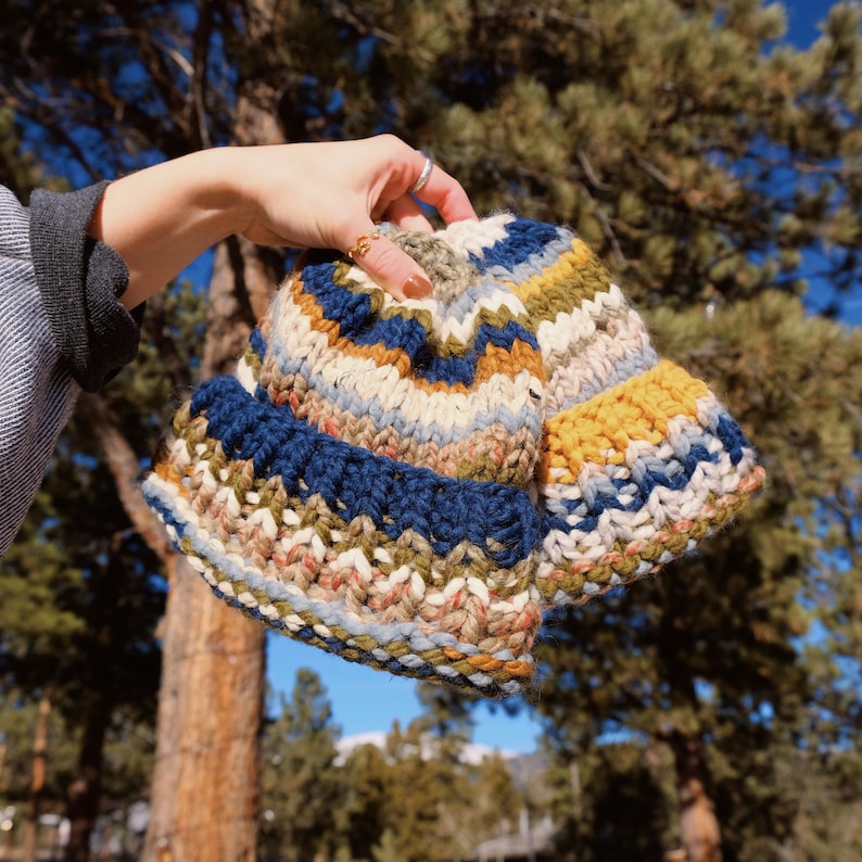 May include: Two knitted hats in a colorful striped pattern with shades of blue, yellow, white, and brown. The hats are held in a person's hand.