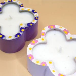 May include: Two clover-shaped candles with white wax and a floral design around the edges. One candle is purple with blue, yellow, and white accents, and the other is light purple with pink and yellow accents.