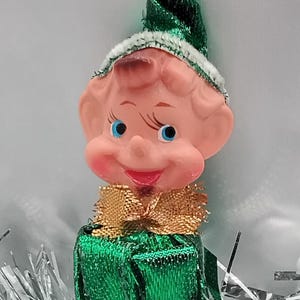May include: A vintage Christmas elf figurine with a pink face, blue eyes, and a smiling red mouth. The elf wears a green outfit, a green hat, and a gold-colored collar. Silver tinsel surrounds the elf.