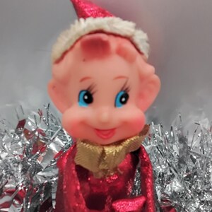May include: A vintage Christmas elf figurine with a red sequined outfit and hat, a gold collar, and a fluffy white trim. The elf has rosy cheeks, blue eyes, and is holding a small, fluffy gift. Silver tinsel is in the background.