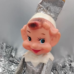May include: A vintage Christmas elf doll with a silver metallic outfit and hat. The elf has a pink face, blue eyes, and a red mouth. The doll is sitting in front of silver tinsel.