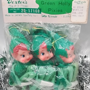 May include: A package of three vintage "Green Holly Pixies" Christmas ornaments. The elves have green felt outfits, red cheeks, and blue eyes. The packaging includes the text "Dexter's" and "LeeWards".