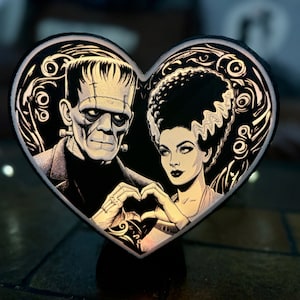 May include: A black and white heart-shaped graphic with a black and white illustration of Frankenstein's monster and his bride. The monster is on the left side of the heart and his bride is on the right side of the heart. They are both making a heart shape with their hands.