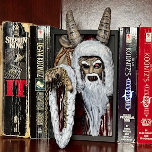 May include: A creepy, white-bearded, red-eyed Santa Claus bookend with horns, holding a gold chain. The bookend is placed between two books with the titles "Dean Koontz's Frankenstein" and "Dean Koontz's Frankenstein".