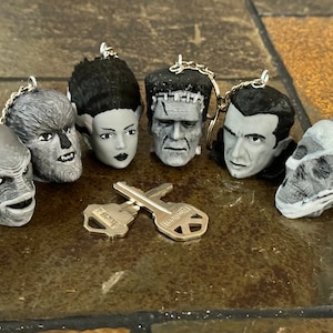 May include: A set of six black and white monster head keychains. The keychains feature the Creature from the Black Lagoon, Frankenstein's Monster, Dracula, the Wolfman, the Mummy, and Bride of Frankenstein.