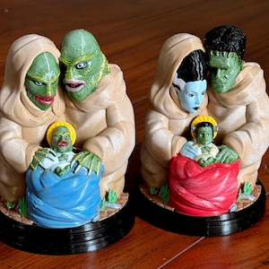 May include: Two monster family figurines. One features a green creature with a beige hooded robe, embracing a smaller green creature in a blue blanket. The other features a green monster with black hair and a blue-skinned monster, embracing a smaller green creature in a red blanket.