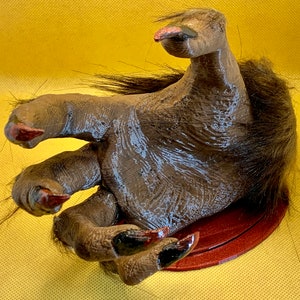 May include: A brown, furry, werewolf hand with long, sharp claws. The hand is posed in a grasping motion and is mounted on a red circular base.