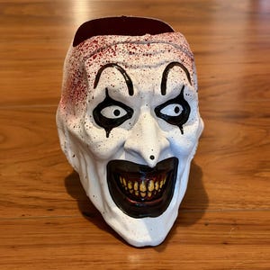May include: A white, open-topped, clown-faced container with black and red accents. The container has a wide, toothy grin, black eye makeup, and red splatters on the top. The container is sitting on a wooden surface.
