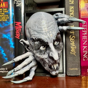 May include: A gray and white bookend shaped like a monster with sharp claws and fangs. The bookend is holding up two books, one titled "Misery" and the other titled "It" by Stephen King.