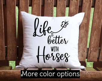 Pode incluir: Travesseiro branco com texto preto que diz "Life is better with horses" e um pequeno gráfico de borboleta. O travesseiro está sentado em um banco de madeira.