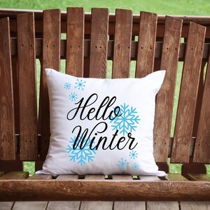 Pode incluir: Almofada quadrada branca com a frase "Hello Winter" em preto e flocos de neve azul claro. A almofada est&aacute; sobre um banco de madeira, com um fundo verde.
