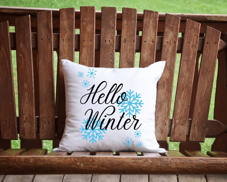 Pode incluir: Almofada quadrada branca com a frase "Hello Winter" em preto e flocos de neve azul claro. A almofada est&aacute; sobre um banco de madeira, com um fundo verde.