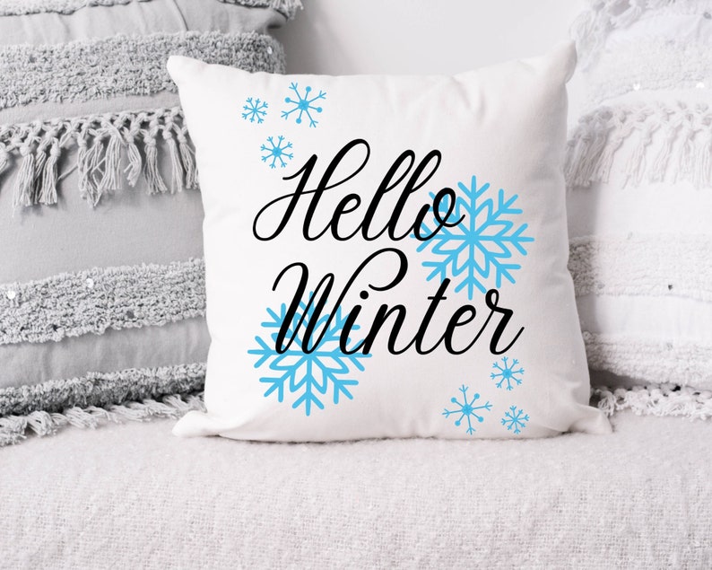 Pode incluir: Almofada quadrada branca com as palavras "Hello Winter" em escrita preta, acompanhada por desenhos de flocos de neve azul claro. A almofada est&aacute; sobre uma superf&iacute;cie branca, com outras almofadas decorativas no fundo.