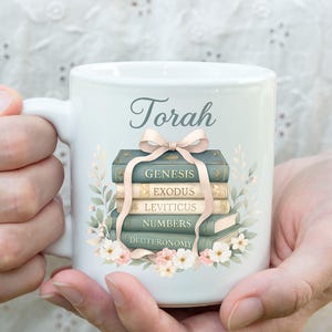 May include: White ceramic mug with the word "Torah" in script. The mug features a stack of books with titles like "Genesis" and "Exodus," tied with a pink ribbon and surrounded by floral accents. A perfect gift for religious studies.