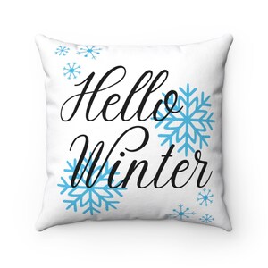 Pode incluir: Almofada quadrada branca com a frase "Hello Winter" em escrita preta, rodeada por desenhos de flocos de neve azul claro. Um elemento decorativo para a casa no inverno.