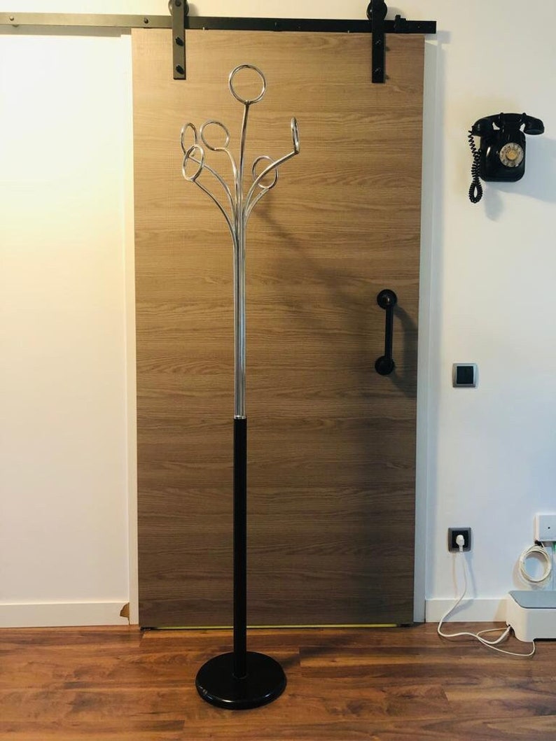 Peut inclure: Un porte-manteau noir et argent avec une base ronde et plusieurs crochets. Le porte-manteau est plac&eacute; devant une porte en bois.