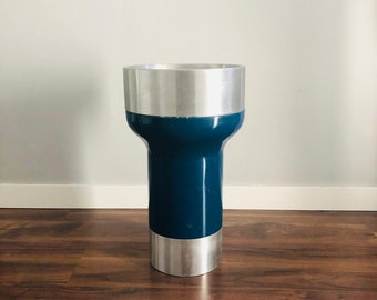 May include: A tall, cylindrical vase with a silver top and bottom and a teal blue middle section. The vase is sitting on a wooden floor.