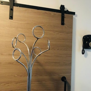 Peut inclure: Un porte-manteau en m&eacute;tal argent&eacute; avec plusieurs crochets en forme d'arbre. Le porte-manteau est plac&eacute; devant une porte coulissante en bois avec des ferrures noires. Un t&eacute;l&eacute;phone rotatif noir est mont&eacute; sur le mur &agrave; droite du porte-manteau.