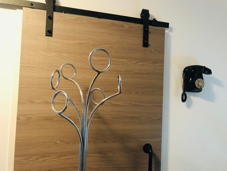 Peut inclure: Un porte-manteau en m&eacute;tal argent&eacute; avec plusieurs crochets en forme d'arbre. Le porte-manteau est plac&eacute; devant une porte coulissante en bois avec des ferrures noires. Un t&eacute;l&eacute;phone rotatif noir est mont&eacute; sur le mur &agrave; droite du porte-manteau.