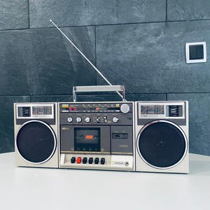 Könnte beinhalten: Ein silber-schwarzer Samsung-Boombox-Player im Vintage-Stil mit ausziehbarer Antenne, zwei großen Lautsprechern und einem Kassettenrekorder. Die Boombox hat einen Tragegriff und verschiedene Bedienelemente.