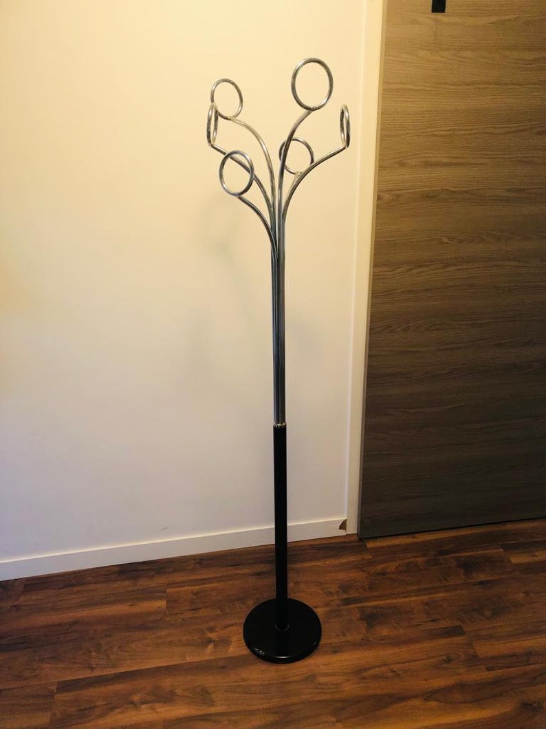 Peut inclure: Un porte-manteau noir et chrom&eacute; avec une base ronde et cinq crochets incurv&eacute;s. Le porte-manteau est pos&eacute; sur un sol en bois.