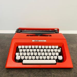 Peut inclure: Une machine à écrire Olivetti Lettera 32 rouge avec un clavier blanc et des touches noires. La machine à écrire porte l'inscription "olivetti COLLEGE" sur le dessus.