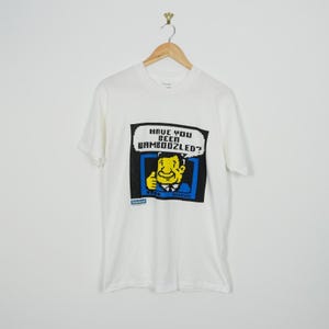 May include: White t-shirt featuring a pixelated graphic with the text "HAVE YOU BEEN BAMBOOZLED?". The graphic shows a smiling cartoon man giving a thumbs up. The t-shirt is displayed on a wooden hanger.