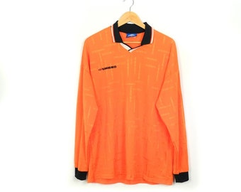 May include: Orange long-sleeved shirt with a black collar and black trim on the sleeves. The shirt has a white v-neck and a small black logo that says "UMBRO" on the left chest.