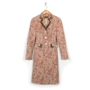 Op de afbeelding: Een lange jas met een bloemenpatroon in roze, beige en bruin, hangend aan een houten hanger. De jas heeft een fluwelen kraag en zakafwerking, met drie witte knopen. Het bloemmotief is gedetailleerd en bedekt de hele jas.