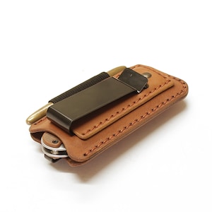 May include: A brown leather wallet with a black metal clip and a small pocket knife. The wallet has a stitched edge and is designed to be carried in a pocket.