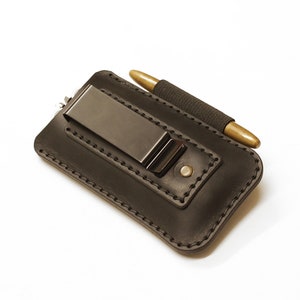 May include: A black leather pen holder with a metal clip and a brass pen. The holder has a stitched border and a small metal rivet. The pen is secured with a black elastic band.