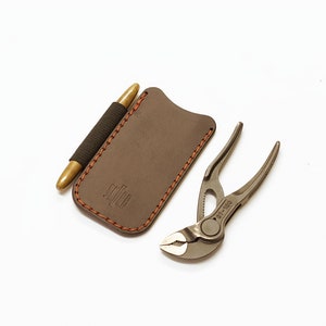 Könnte beinhalten: Eine braune Ledertasche mit orangefarbenen Nähten und einem Stifthalter. Eine silberne Multifunktionszange mit dem Text "DT-100" liegt neben der Tasche.