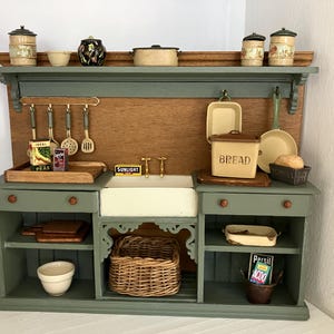 May include: A miniature sage green kitchen unit with a wooden backsplash, featuring a central white sink, a wicker basket, open shelving, and various small kitchen accessories including a bread bin, pots, pans, and miniature food boxes.